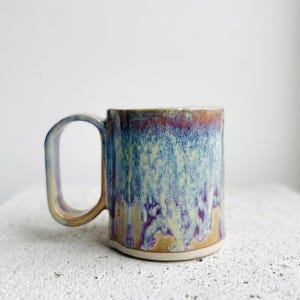 May include: A ceramic mug featuring a reactive glaze with a blend of blue, purple, and tan colors. The glaze exhibits a dripped, layered effect across the cylindrical body. It includes a large, angular handle attached to the side of the vessel.