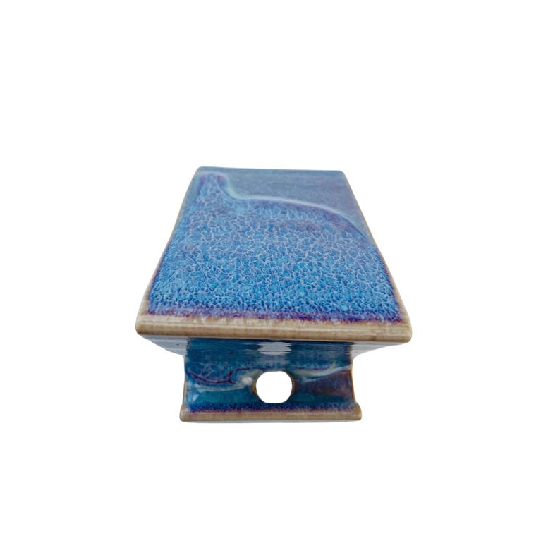 May include: A rectangular ceramic object with a textured, mottled blue and purple glaze on the top surface. The base features a smooth, glossy finish in shades of blue and brown, with a single circular hole centered on the front-facing side.