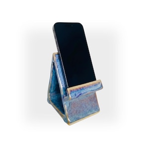 May include: A triangular ceramic phone stand with a glossy, mottled blue and purple glaze finish. The stand features a wide base and a front ledge designed to hold a black smartphone in an upright position, showcasing a smooth, reflective surface.