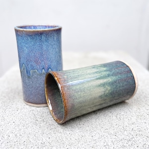 May include: Two cylindrical ceramic tumblers with smooth, glazed surfaces. One tumbler features a vibrant blue and purple mottled glaze with a drip pattern, while the other displays a gradient of sage green and brown tones with a glossy finish.