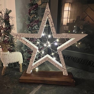 May include: A large, white, star-shaped Christmas decoration with a distressed finish. The star is illuminated by small white lights and sits on a dark wooden base. A decorative reindeer and a small Christmas tree are in the background.