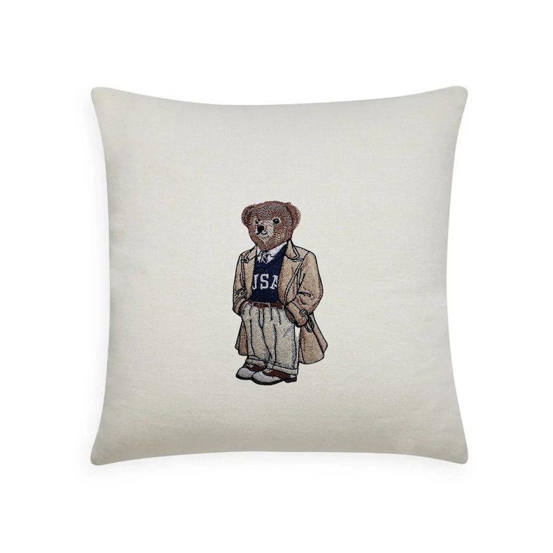 May include: Cream-colored square pillow featuring an embroidered bear wearing a tan overcoat, a blue sweater with "USA" on it, and gray pants. The bear is standing upright. The pillow is a decorative item.