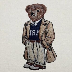 May include: Embroidered patch featuring a brown teddy bear wearing a tan trench coat, navy blue sweater with "USA" text, and khaki pants. The bear has a detailed face and is wearing a tie. The patch is designed with a classic, preppy style.