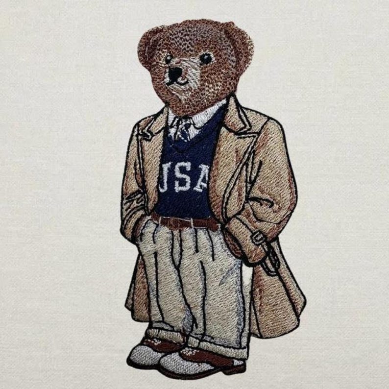 May include: Embroidered patch featuring a brown teddy bear wearing a tan trench coat, navy blue sweater with "USA" text, and khaki pants. The bear has a detailed face and is wearing a tie. The patch is designed with a classic, preppy style.
