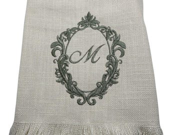 May include: Off-white linen towel with a decorative embroidered oval frame in a muted green color. The frame surrounds the letter "M". The bottom edge of the towel has a fringe.