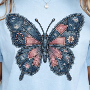 May include: Pale blue t-shirt with a large butterfly design. The butterfly is made from denim patches in blue and pink, with distressed details. A small flower and floral elements are incorporated into the design. The shirt has a round neckline.