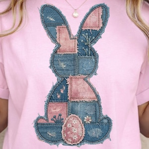 May include: A light pink t-shirt featuring a patchwork denim bunny design with pink accents and a floral egg. The bunny has frayed edges, giving it a rustic look. The design is centered on the shirt.