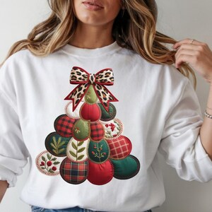 May include: White sweatshirt featuring a Christmas tree design. The tree is composed of red, green, and plaid fabric circles, topped with a leopard print bow. The design has a stitched, quilted appearance.