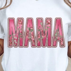 Peut inclure: T-shirt blanc avec le mot "MAMA" en grandes lettres roses. Chaque lettre est ornée d'un motif floral et bordée de perles. Le t-shirt est de style décontracté.
