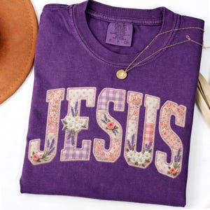 Könnte beinhalten: Ein lila Comfort Colors T-Shirt mit dem Wort "JESUS" in einem Patchwork-Design appliziert. Die Buchstaben zeigen florale und gemusterte Stoffe, darunter Lavendel, Gänseblümchen und rosa Rosen. Eine goldene Halskette mit Anhänger liegt auf dem Shirt.