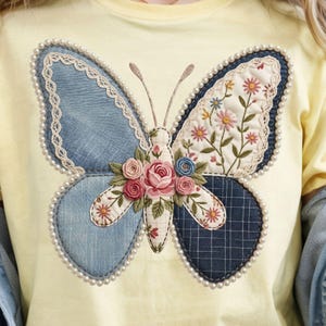 May include: A yellow t-shirt featuring a large butterfly applique. The butterfly has denim and floral patterned wings, embellished with pearls and lace. The butterfly's body is adorned with fabric roses and floral details.