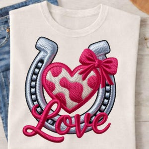Puede incluir: Camiseta blanca con un diseño bordado que presenta una herradura plateada, un corazón rosa con lunares blancos y un lazo rosa. La palabra "Love" está bordada en rosa debajo.