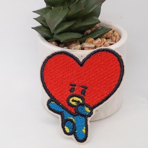 Puede incluir: Parche bordado con un personaje rojo en forma de corazón con un contorno negro. El personaje tiene brazos azules y detalles amarillos. El parche está delante de una pequeña maceta de cerámica blanca con una planta suculenta verde.