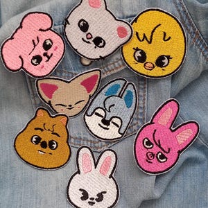 May include: A selection of embroidered patches depicting cartoon animal faces. The patches include a pink dog, a grey bear, a yellow chick, a tan fox, a blue bunny, a brown cat, a white bunny, and a pink bunny with an angry expression. The patches are on a denim background.