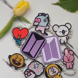 Op de afbeelding: Een verzameling kleurrijke geborduurde patches met stripfiguren en een paars logo. De patches zijn gerangschikt op een wit oppervlak met een gele tulp en een parelketting. De patches zijn in verschillende vormen en maten.