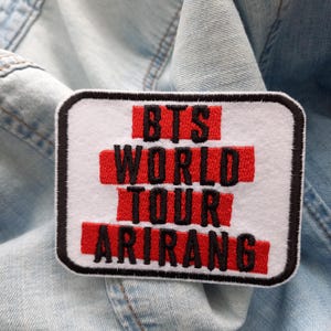 Könnte beinhalten: Ein rechteckiger Aufnäher mit dem Text "BTS WORLD TOUR ARIRANG" in Rot und Schwarz auf weißem Grund. Der Aufnäher hat einen schwarzen Rand und liegt auf einem hellblauen Jeansstoff.