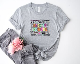 May include: A grey t-shirt featuring a colourful "Back to School" design with words like "Homework" and "Crayons". The shirt is styled with grey jeans and a pink flower. The design is suitable for the school season.