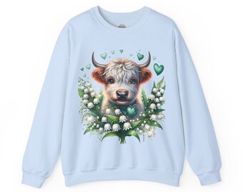 May include: Pale blue sweatshirt with a Highland cow graphic. The cow has white fur, brown ears, and a green heart-shaped collar. It's surrounded by white lily of the valley flowers and green heart accents. A whimsical and charming design.