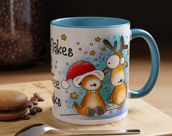 May include: White ceramic mug with a blue interior and handle. The mug features a whimsical illustration of two cartoon characters, one wearing a Santa hat, with the text "Snowflakes make coffee breaks". A spoon and a macaron are on the wooden surface.
