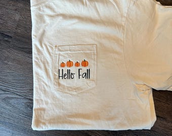 Może przedstawiać: Kremowa koszulka z kieszonką z trzema pomarańczowymi dyniami i czarnym napisem "Hello Fall". Koszulka jest złożona na ciemnej drewnianej powierzchni.