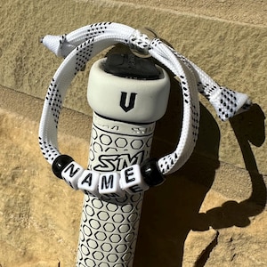 May include: A hockey stick with a white and black patterned grip. A white bracelet with black letters spelling "NAME" is attached. The stick's end has a white top with a black "U" and the text "SM ME".