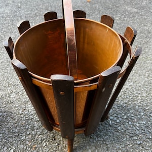 May include: A copper plant stand with a wooden frame and a handle. The stand has a cylindrical shape and three legs.