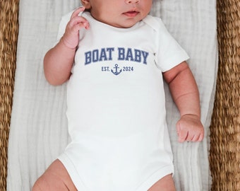 May include: A white baby onesie with blue text that reads "BOAT BABY EST. 2024" and a small anchor symbol.
