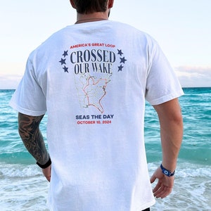 May include: White t-shirt with a red, white, and blue graphic of a map of the United States. The graphic says "America's Great Loop Crossed Our Wake" and "Seas the Day October 10, 2024".