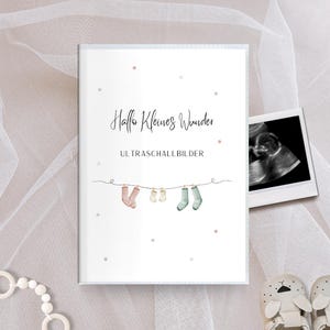 Könnte beinhalten: Eine weiße Karte mit dem Text "Hallo Kleines Wunder" und "Ultraschallbilder" mit einer Reihe von Babysocken, die daran hängen.
