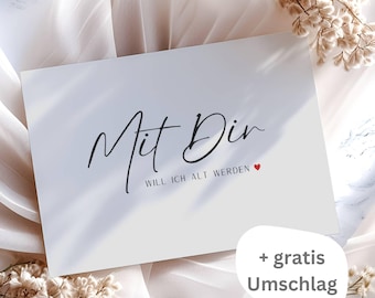 May include: A white card with the text "Mit Dir Will Ich Alt Werden" in black script with a small red heart. The card is on a white fabric background with dried flowers. The text "+ gratis Umschlag" is in black text on a white background.