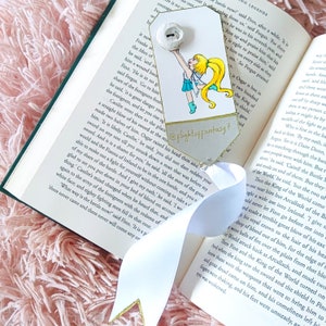 Puede incluir: Un marcapáginas blanco con un diseño dorado y una ilustración de dibujos animados de una niña sosteniendo una espada. El marcapáginas tiene el texto "@Flightoffantasy7" impreso en él.