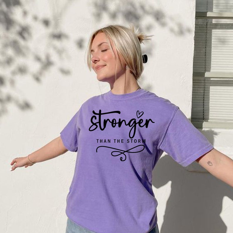 May include: A purple t-shirt with the text "stronger than the storm" and a small heart graphic.