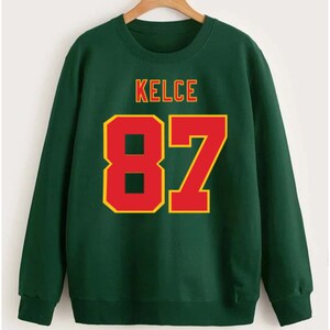 May include: Green sweatshirt with the name "KELCE" and the number "87" printed in red and yellow.