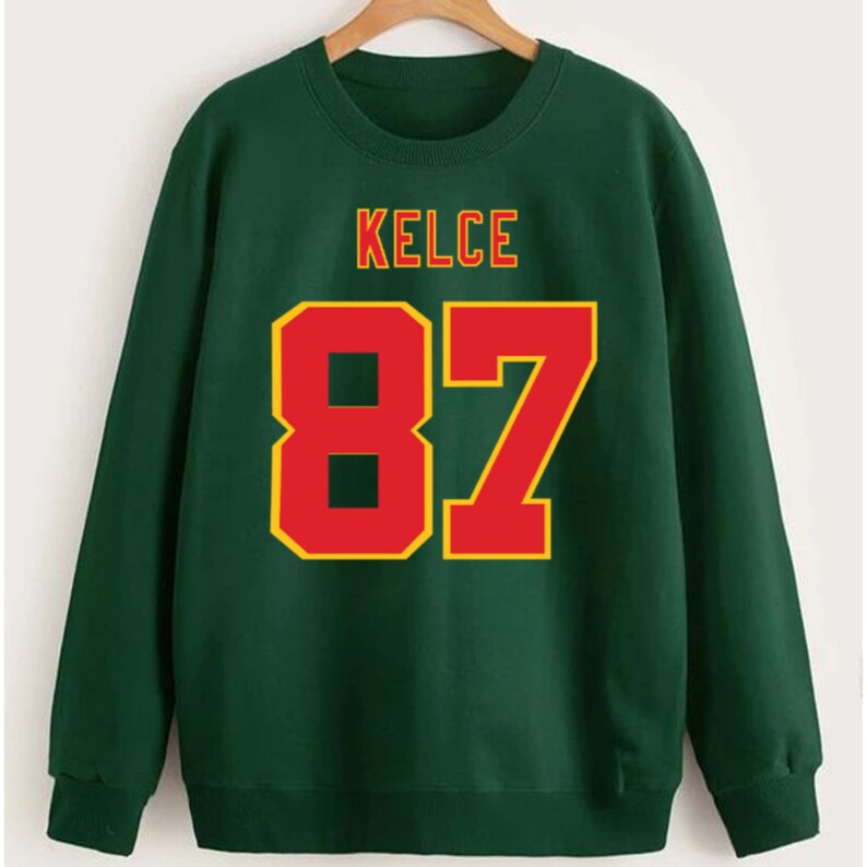 May include: Green sweatshirt with the name "KELCE" and the number "87" printed in red and yellow.