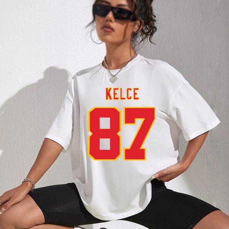 May include: White oversized t-shirt with red and yellow lettering that says "KELCE 87".