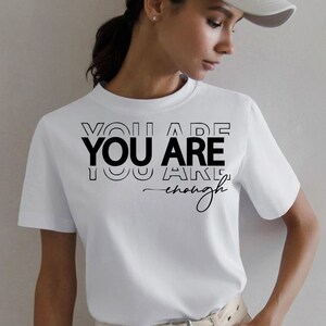 May include: White t-shirt with black text that reads "You Are You Are You Are enough" in a layered, stacked font.