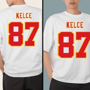 May include: White t-shirt with red and yellow lettering that says "KELCE 87".