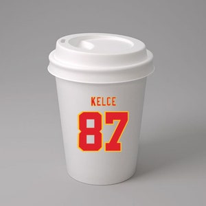 May include: White to-go coffee cup with a red and yellow design. The cup has the name "KELCE" and the number "87" printed on it.