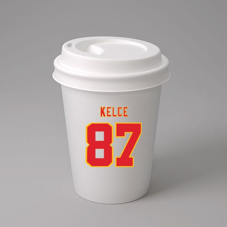 May include: White to-go coffee cup with a red and yellow design. The cup has the name "KELCE" and the number "87" printed on it.