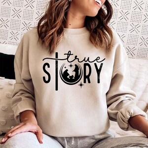 May include: A beige sweatshirt with a black graphic design that says "true story" with a depiction of the nativity scene inside a crescent moon.