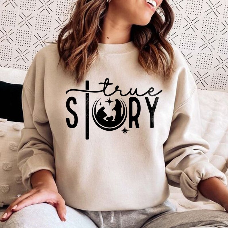 May include: A beige sweatshirt with a black graphic design that says "true story" with a depiction of the nativity scene inside a crescent moon.