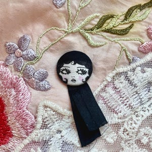 Könnte beinhalten: Ein runder, handbemalter Knopf mit einem Vintage-Puppengesicht-Design. Der Knopf hat eine schwarze Bob-Frisur, zarte Gesichtszüge und ein schwarzes Band. Er ist vor einem Hintergrund aus besticktem Stoff mit floralen und Spitzen Details platziert.