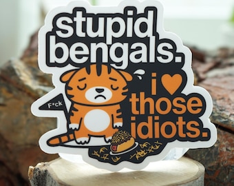 Pode incluir: Um autocolante com o texto "stupid bengals. i love those idiots." apresenta um tigre de desenho animado com uma expressão triste. O tigre é laranja e branco com listras pretas, segurando uma pequena bandeira que diz "f*ck".