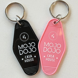Puede incluir: Dos llaveros negros y rosas con un diseño de etiqueta de llave de motel. Los llaveros presentan el texto "MOJO DOJO CASA HOUSE" en letras blancas.