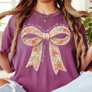May include: Purple t-shirt featuring a large bow design with floral accents. The bow is cream-colored with pink, yellow, and green flowers. The text "...she is clothed with strength & dignity" and "Proverbs 31:25" are printed on the shirt.