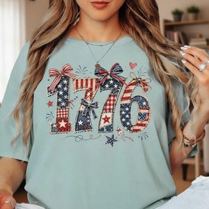 May include: A light blue short-sleeve t-shirt featuring the number 1776 in a red, white, and blue patriotic pattern. The design includes stars, stripes, and plaid textures, accented with decorative bows, small hearts, and star illustrations.