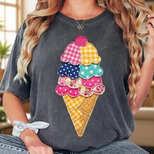 May include: Dark gray t-shirt featuring a colorful ice cream cone design. The ice cream cone is made of various patterned fabrics, including gingham, polka dots, and floral prints. The cone is a waffle pattern. The shirt is a casual, comfortable style.