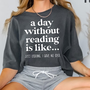 May include: A dark gray t-shirt with the text "a day without reading is like... JUST KIDDING, I HAVE NO IDEA." The shirt is paired with gray shorts. A white vase with decorative grass is in the background.