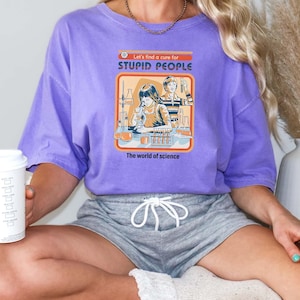 May include: A lilac-colored t-shirt with the text "Let's find a cure for STUPID PEOPLE" and an illustration of scientists in a lab. The words "The world of science" are below the illustration. The shirt is part of the "Trendy Sweats handmade apparel" collection.