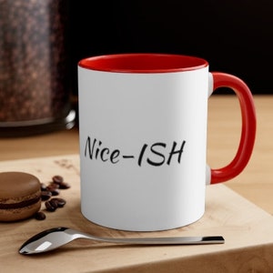 May include: White ceramic coffee mug with a red rim and handle. The mug has the text "Nice-ISH" printed in black on the front.